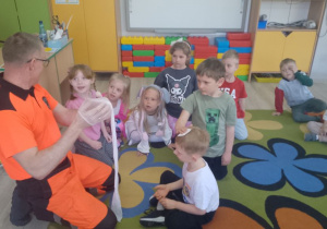 Na środku koła siedzi Bruno z opatrunkiem na głowie, który trzyma Marcel, ratownik trzyma opaskę uciskową. Obok siedzą dzieci.