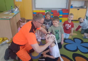 Ratownik zakłada na głowę chłopca opaskę uciskową. Z boku przyglądają się przedszkolaki.