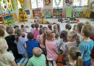 Dzieci siedzą na dywanie, a przed nimi dwóch strażaków demonstruje ułożenie poszkodowanego w pozycji bezpiecznej. W tle kąciki zainteresowań, okna.