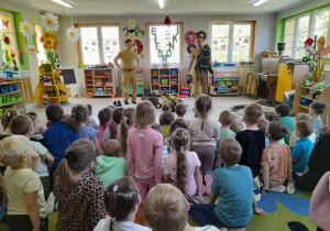 Dzieci siedzą na dywanie, a przed nimi strażak prezentuje aparat powietrzny.