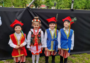 Czworo dzieci z grupy "Słoneczka" stoi obok sceny w parku Traugutta. Przedszkolaki ubrane są w stroje krakowskie.