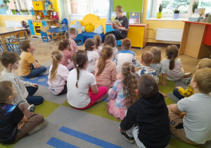 Dzieci siedzą na dywanie, a kolega ze szkoły czyta książkę. W tle tablica, kącik domowy, kanapa.