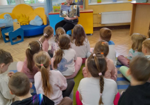 Przedszkolaki siedzą na dywanie, a kolega ze szkoły prezentuje ilustrację w książce.