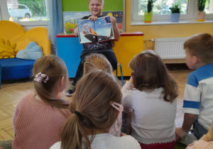 Dzieci siedzą na dywanie, a kolega ze szkoły prezentuje kolejną ilustrację w książce. W tle tablica, okna.