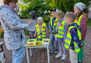 Dzieci stoją obok pomnika dr. A. Troczewskiego, a przed nimi znajduje się stolik z obrazkami ilustrującymi legendy. Pani czyta dzieciom fragmenty legend. W tle Pałac Ślubów, drzewa.
