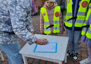 Pani przydziela dzieciom punkty za wykonanie zadania - stempelki z symbolami róży. Obok stoi Antek i Łucja. W tle pomnik, Pałac Ślubów, drzewo.