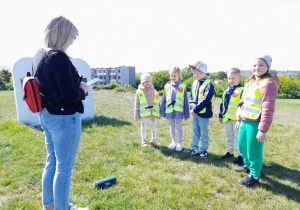 Dzieci stoją na Cmentarzu żydowskim obok pamiątkowej tablicy, a przed nimi nauczyciel z Przedszkola Miejskiego nr 17 "Niezapominajka" w Kutnie. "Słoneczka" rozpoznają ze słuchu hymn Polski.