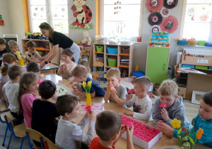 Dzieci siedzą przy wspólnym stole i oglądają, wąchają mydlane kwiaty, które znajdują się w pudełkach. Obok pani Kasia stawia na stole pudełko z kwiatami.