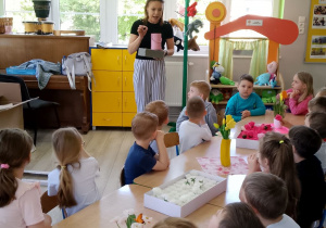 "Biedronki" siedzą przy stole, przed nimi stoi pani Kasia, która trzyma różowe pudełko i gąbkę florystyczną.