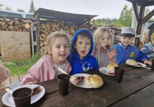 Zuzia, Wojtuś, Nadia i Oluś siedzą przy stole i jedzą kiełbaski.