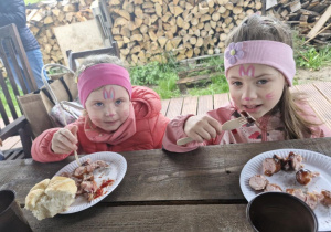 Ola i Oliwka jedzą pieczone na ognisku kiełbaski.