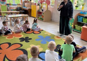 Dzieci siedzą w dwóch rzędach. Pani z biblioteki stoi pomiędzy nimi.