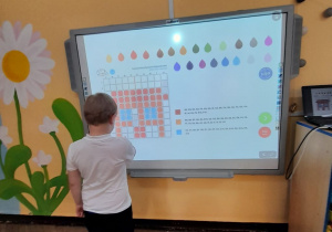 Adam stoi przed tablicą multimedialną i układa obrazek na podstawie podanych koordynatów.