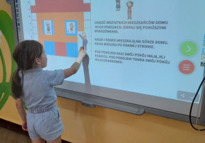 Na tablicy multimedialnej wyświetlony jest domek i jego mieszkańcy. Matylda przesuwa obrazek przedstawiający członka rodziny do właściwego pokoju.