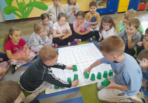 Patryk i Antek układają na dole maty ramkę z zielonych kubków zgodnie ze słowną instrukcją. Koleżanki i koledzy obserwują działanie chłopców.
