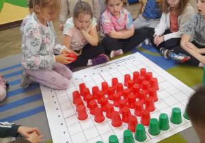 Ula układa na macie do kodowania serce z czerwonych kubków zgodnie z treścią rymowanki. Grupka dzieci obserwuje działanie dziewczynki.