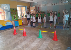 Dzieci ubrane w białe i zielone koszulki obracają się z podniesionymi do góry rękami podczas rozgrzewki.