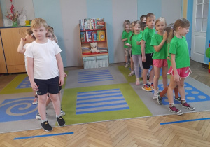 Dzieci ubrane w białe i zielone koszulki stoją w dwóch rzędach przed wyznaczonymi liniami startu.