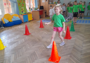 Ula wykonuje slalom z woreczkiem na głowie między pachołkami. W tle pozostałe dzieci czekają na swoją kolej.