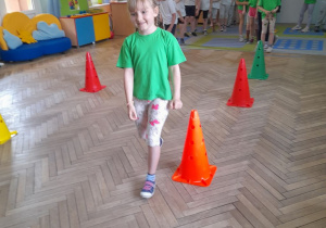 Oliwia wykonuje slalom z woreczkiem na głowie między pachołkami. W tle pozostałe dzieci czekają na swoją kolej.