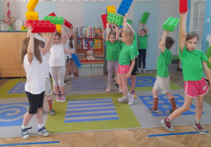 Dzieci stoją na dywanie i podnoszą do góry duży, plastikowy klocek.