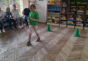 Oliwia przenosi piłeczkę na paletce.W tle szafki z układankami. Z lewej strony siedzą rodzice na krzesełkach.