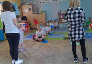 Przedszkolaki siedzą na dywanie w dwóch rzędach, a przed nimi stoją dwie mamy z medalami na tacach.