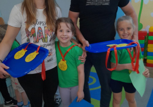 Wiktoria i Julka stoją obok swoich rodziców, którzy trzymają tacki z medalami. Dziewczynki mają zawierzone na szyi medale.