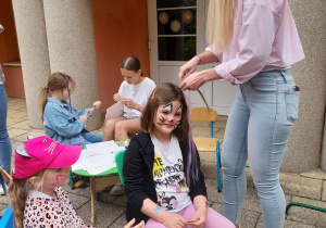 Trzy dziewczynki siedzą na krzesłach podczas zaplatania warkoczyków i malowania brokatowych tatuaży.