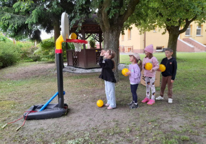 Wiktoria rzuca piłkę do kosza. Za nią stoi troje dzieci. W tle drzewo, altana.