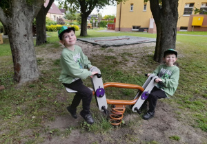 Antek i Franek siedzą na huśtawce. W tle drzewa, piaskownica, fragment budynku przedszkola.