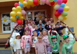 Grupa "Słoneczek" pozuje do zdjęcia przed wejściem do przedszkola. Nad wejściem girlanda z kolorowych balonów i balony w kształcie stokrotek.