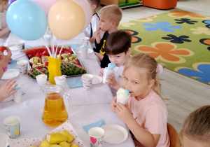 Grupka dzieci siedzi przy wspólnym stole podczas poczęstunku. Przedszkolaki jedzą watę cukrową, owoce i piją sok.