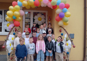 Zdjęcie grupowe dzieci przy wejściu głównym do przedszkola. Wejście udekorowane kolorową girlandą balonową oraz balonami w kształcie stokrotek.