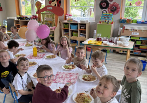 Dzieci siedzą przy stole, przy którym podane jest drugie danie – spaghetti. Dzieci uśmiechają się do zdjęcia.
