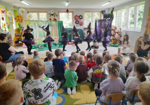 Sześć dziewczynek w roli Czarownic porusza się przy piosence "Kochajcie Czarownice". Dzieci oglądają występ rówieśników.