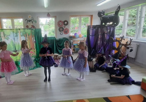 Na scenie stoi Mała Czarowniczka, a obok pięć dziewczynek w roli Wróżek. Z boku siedzi Julka w roli Narratora. W tle chata Czarownic i dekoracja lasu.