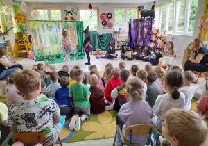 Dzieci oglądają przedstawienie pt. "Mała Czarowniczka". Na środku sceny Nikola w roli Myszki i Ula w roli Małej Czarownicy odgrywają swoje role. W tle dekoracja lasu i chata Czarownic.