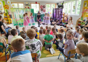 Przedszkolaki z zainteresowaniem oglądają przedstawienie w wykonaniu swoich kolegów. Mała Czarowniczka stoi na środku sceny, a obok niej pięć Wróżek. W tle dekoracja lasu oraz chata Czarownic.