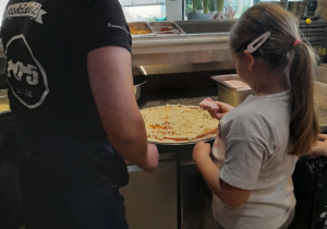 Lena rozkłada na okrągłym placku pizzy szynkę. Obok stoi Pani pracująca w pizzerii.