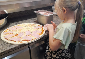 Oliwia rozkłada na okrągłym placku pizzy szynkę.
