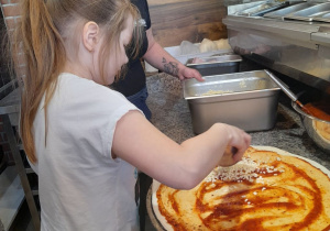 Julka pod okiem Pani z pizzerii rozkłada ser na placek polany sosem pomidorowym.