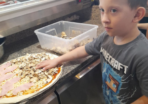 Wojtek rozkłada na okrągłym placku pizzy pieczarki.