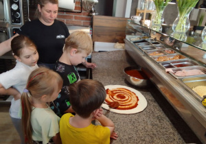Pod okiem Pani z pizzerii Adam smaruje placek sosem pomidorowym. Obok Lena, Oliwia i Antek obserwują działania Adama.