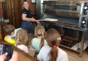 Dzieci obserwują, jak pracownica pizzerii wkłada pizzę do pieca.