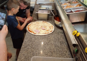 Mikołaj rozkłada na okrągłym placku pizzy szynkę.