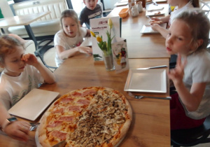 Grupa dzieci przy stole, na którym są talerze, sztućce oraz ogromna pizza na okrągłym talerzu.