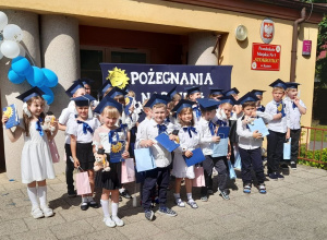 Dzieci z dyplomami i upominkami pozują do wspólnego zdjęcia na tarasie. Przedszkolaki ubrane są na galowo, na głowach mają birety.