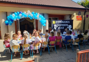 Dzieci siedzą przy połączonych stołach. W tle budynek przedszkola i dekoracja: nad drzwiami wisi biało-niebieska girlanda z balonów, obok na granatowym materiale przypięte są dwie postacie absolwentów przedszkola, słoneczko i napis: "Pożegnania nadszedł czas".