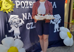 Mama naszej wychowanki stoi na tle dekoracji i trzyma tacę z tortem.